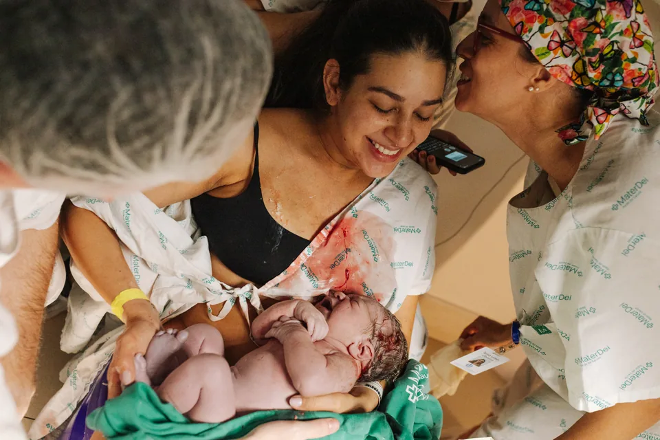 Mãe sorri com o bebê no colo após o nascimento, acompanhada pela equipe